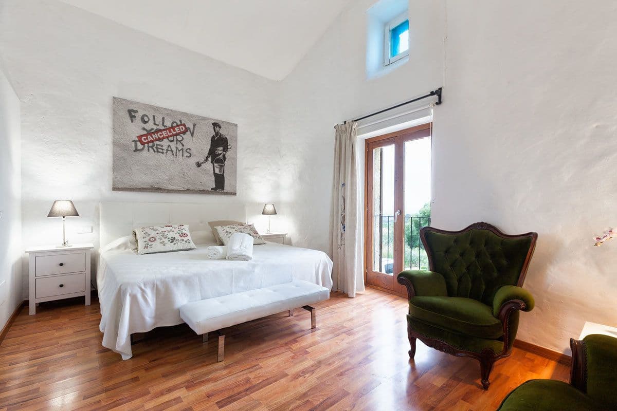 Un dormitorio con una cama, dos mesitas de noche, un sillón verde y una puerta al balcón.