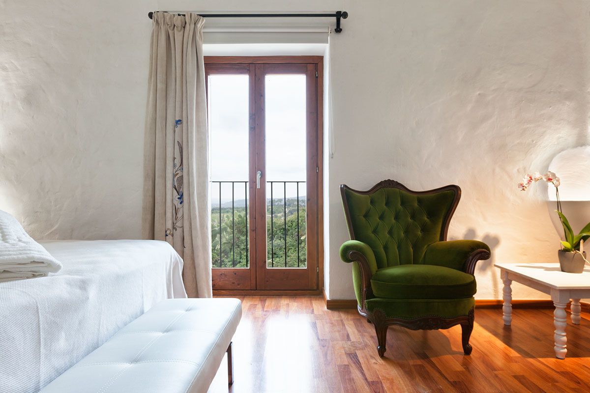 A bedroom with a bed, a green armchair, a small table with a plant, and a view through a door.