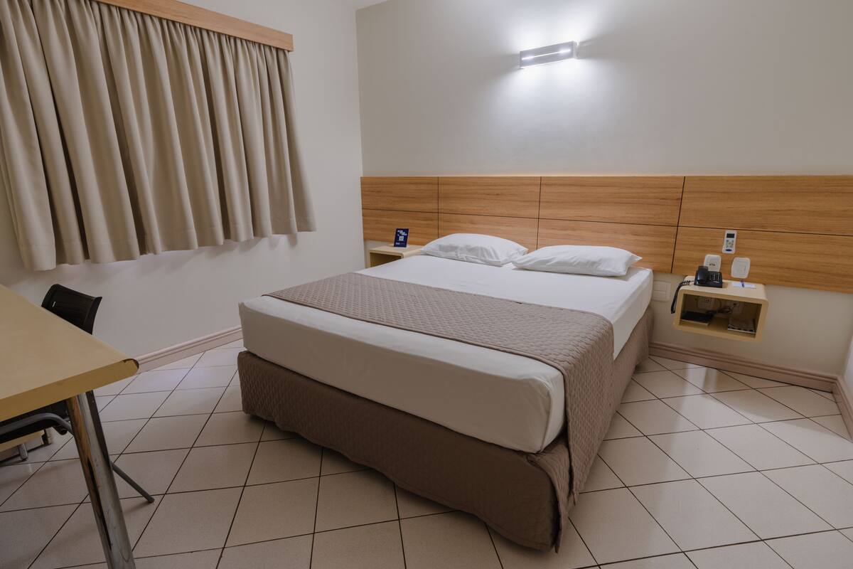 Una habitación de hotel con una cama grande, una mesita de noche, un escritorio y una ventana con cortinas.