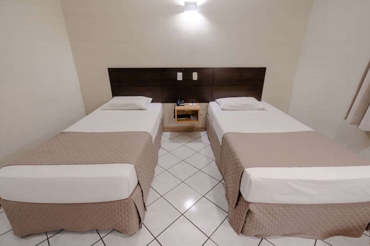 Habitación de hotel con dos camas individuales, una mesita de noche de madera y piso de baldosas.