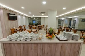 Un área de bufé con varias mesas y sillas para comer, un televisor montado en la pared, y una variedad de alimentos y bebidas exhibidos en la barra.