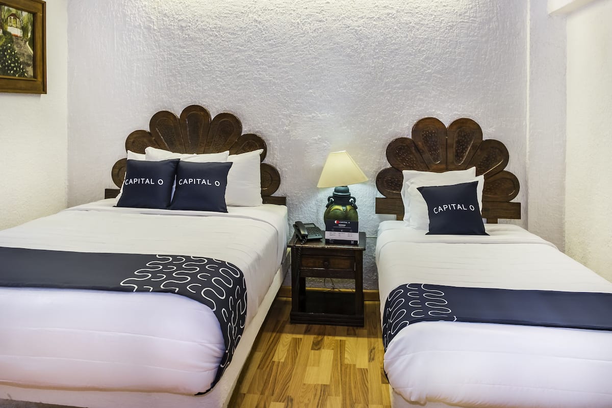 Habitación de hotel con dos camas, cada una con un diseño único de cabecera de madera, ropa de cama blanca y almohadas a juego. Un mesita de noche con una lámpara y un teléfono está entre las camas.