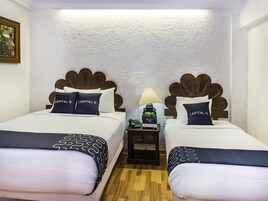 Habitación de hotel con dos camas, cada una con un diseño único de cabecera de madera, ropa de cama blanca y almohadas a juego. Un mesita de noche con una lámpara y un teléfono está entre las camas.