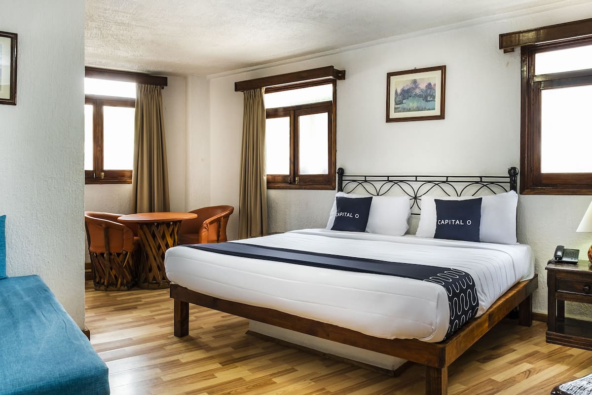 Habitación de hotel con una cama de madera, una mesita con sillas y un cuadro en la pared.