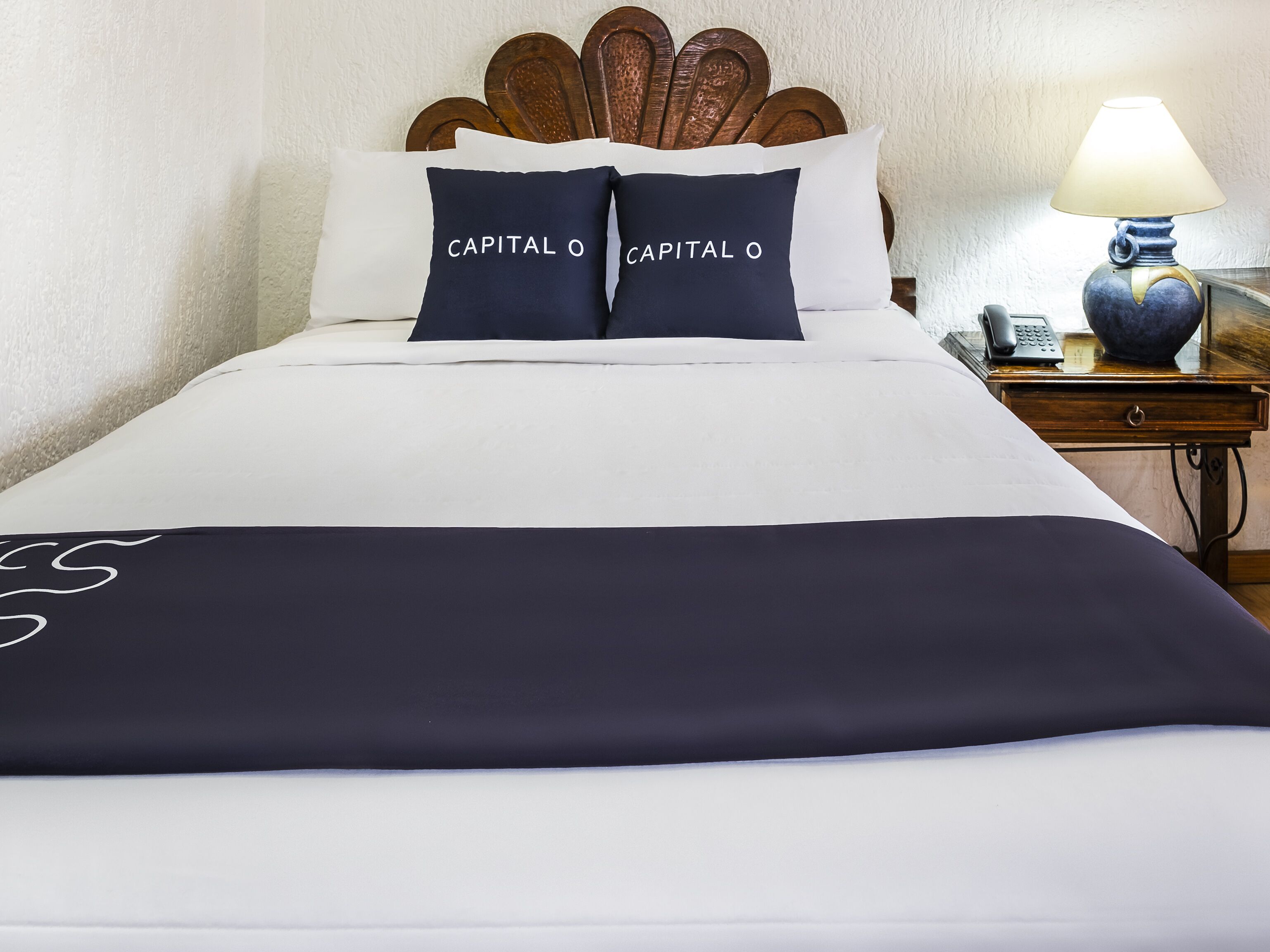 Una cama bien hecha con colcha blanco y azul marino, dos almohadas azul marino con el texto 'CAPITAL O', un cabecero de madera, una mesita de noche con lámpara y teléfono.