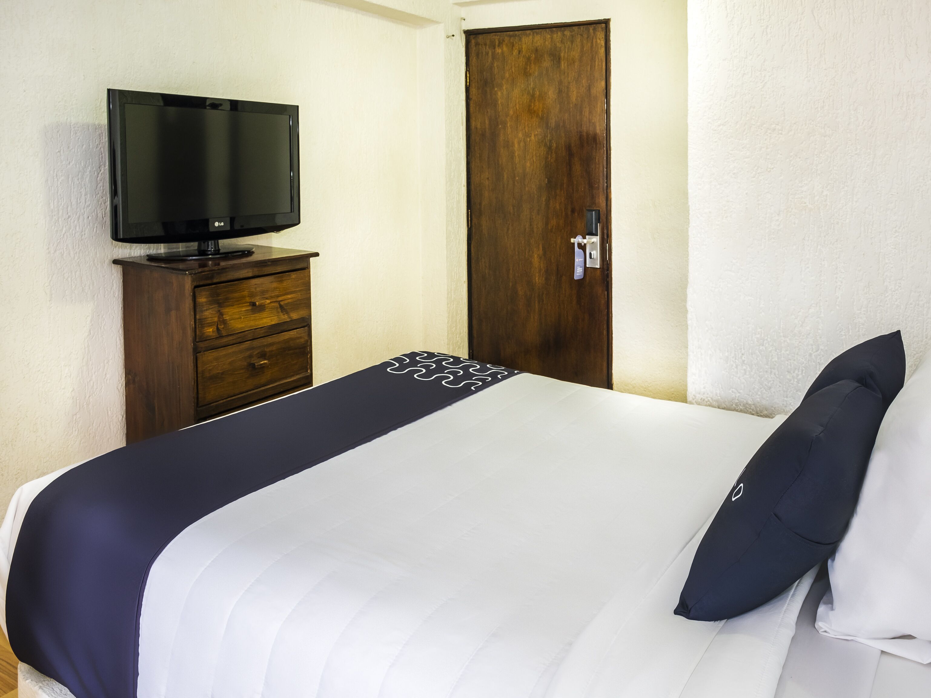 A hotel room with a bed, a TV on a wooden dresser, and a wooden door.