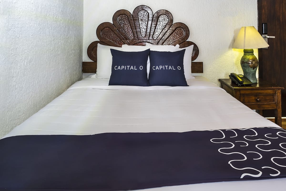Una cama bien hecha con ropa blanca y dos almohadas azul marino con el texto "CAPITAL O", un cabecero de madera con un diseño de hojas, una lámpara de noche y una mesita de noche de madera con un teléfono.