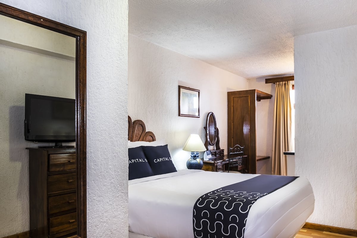Habitación de hotel con una cama, televisión, cómoda, espejo y ventana con cortinas.