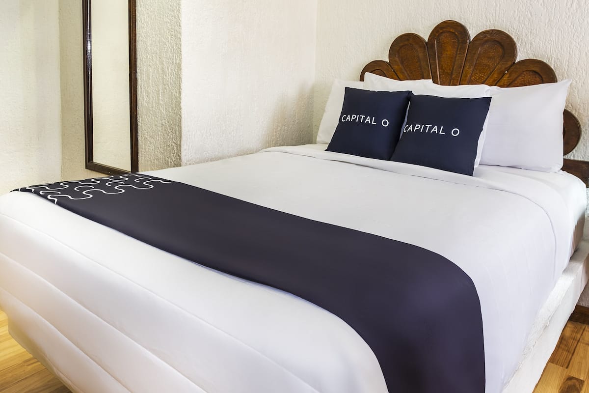 Una cama bien hecha con ropa de cama blanca y azul marino, dos almohadas azul marino con el texto 'CAPITAL O' y un cabecero de madera.