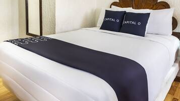 Una cama bien hecha con ropa de cama blanca y azul marino, dos almohadas azul marino con el texto 'CAPITAL O' y un cabecero de madera.