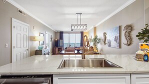 A modern kitchen with a stainless steel sink, white cabinetry, and a marble countertop. There is a dining area with a view of the ocean in the background.