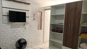 Une pièce avec un téléviseur fixé au mur, qui est fait de briques blanches, un ventilateur sur pied, une armoire avec des étagères et un tapis à motifs.
