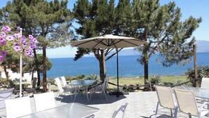 Un área de estar al aire libre con mesas y sillas, una pérgola con sombrilla, y vista al mar.