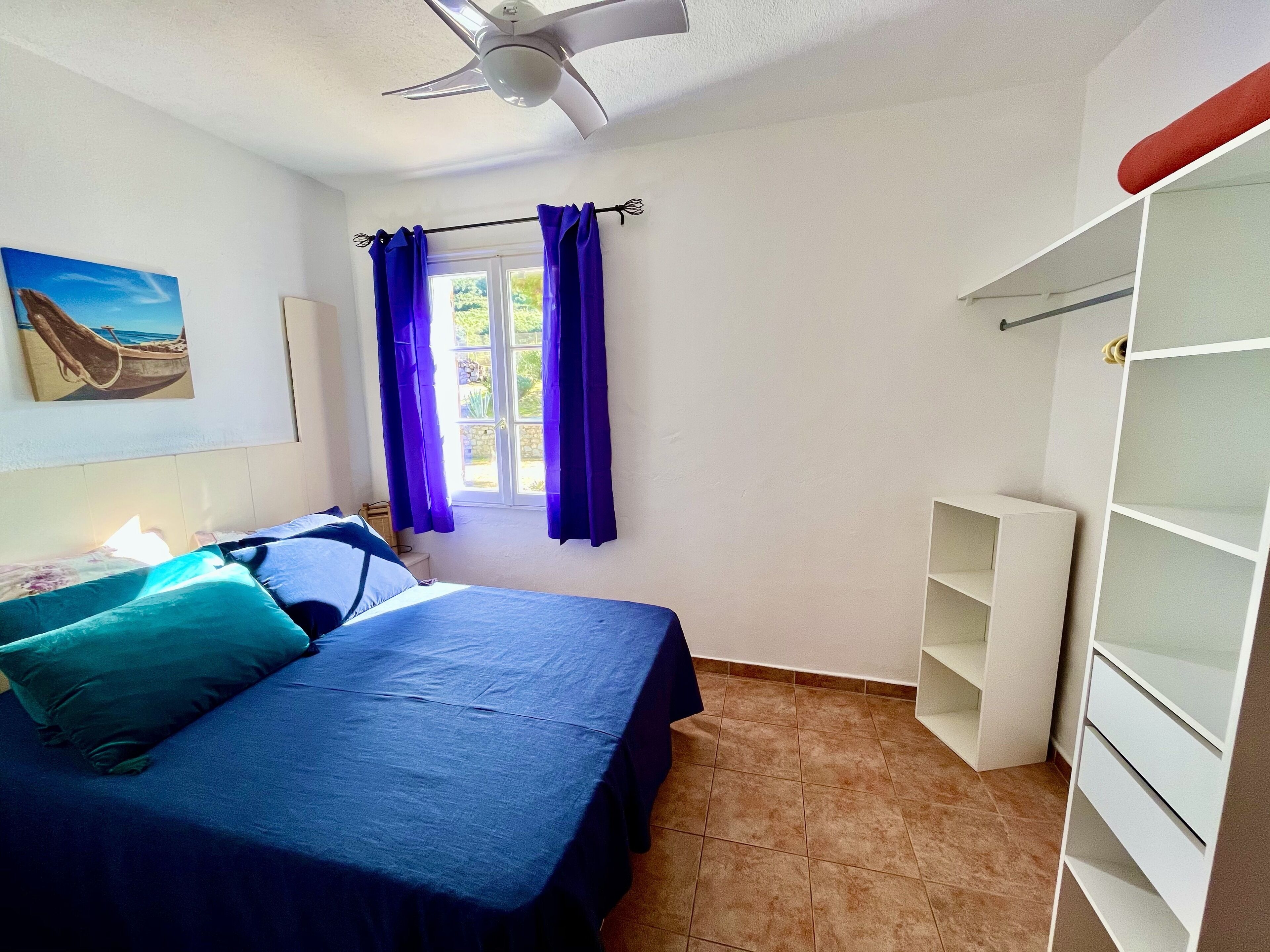 A bedroom with a bed, a ceiling fan, a window with purple curtains, a painting on the wall, and a white wardrobe.