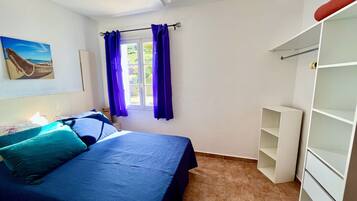 A bedroom with a bed, a ceiling fan, a window with purple curtains, a painting on the wall, and a white wardrobe.