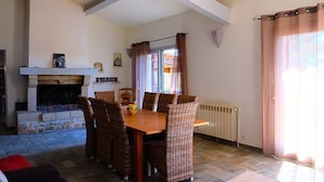 A dining area with a stone fireplace, wicker chairs, a wooden table, and a view through a window with curtains.