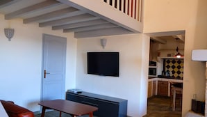 A living room with a TV, a sofa, a table, and a kitchen area visible through an open doorway.
