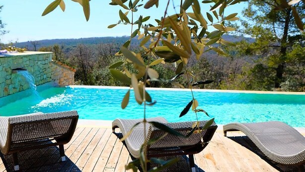A wooden deck with lounge chairs overlooking a swimming pool with a waterfall feature, set against a backdrop of trees and hills.