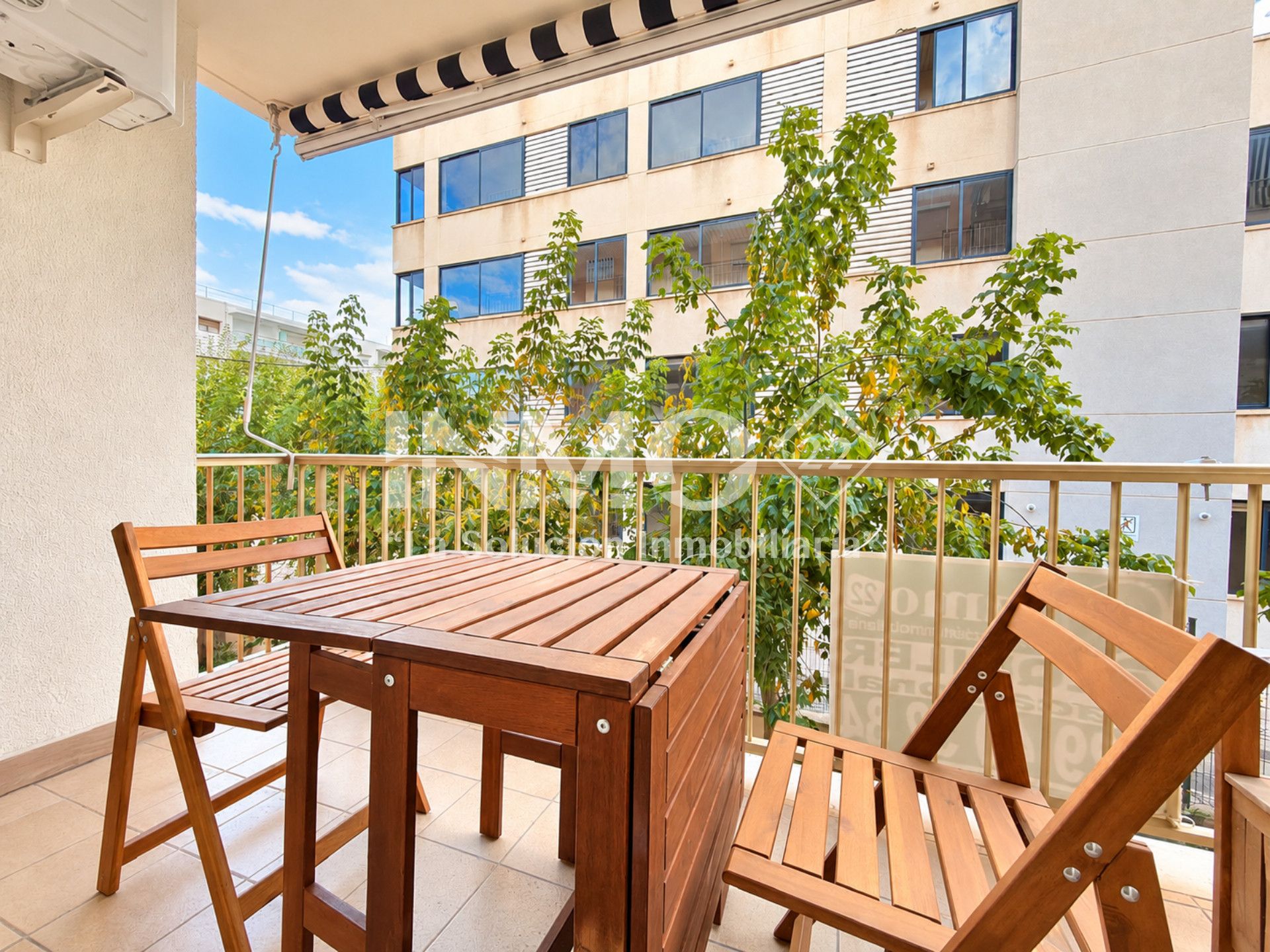 Un balcón con mesa y sillas de madera, barandilla y vista a un edificio con ventanas y vegetación.