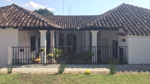 A two-story building with a tiled roof, a covered patio with a railing, and a small garden area with plants. - Casa LOBE (Ocosingo)