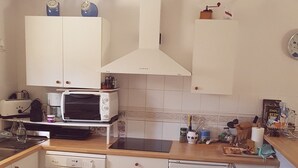Een functionele keuken met witte kastjes, een magnetron, een broodrooster en een vaatwasser. Twee barkrukken en een klein spoelbakje zijn aanwezig.