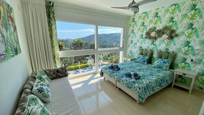 Une chambre avec une grande fenêtre, un lit recouvert d’une couvre-lit à motifs de feuilles, un ventilateur de plafond et une vue sur l’extérieur.