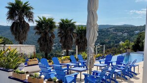 Un espace aménagé au bord de la piscine, avec des chaises et des tables bleues, entouré de plantes et de palmiers, et offrant une vue sur un paysage pittoresque.