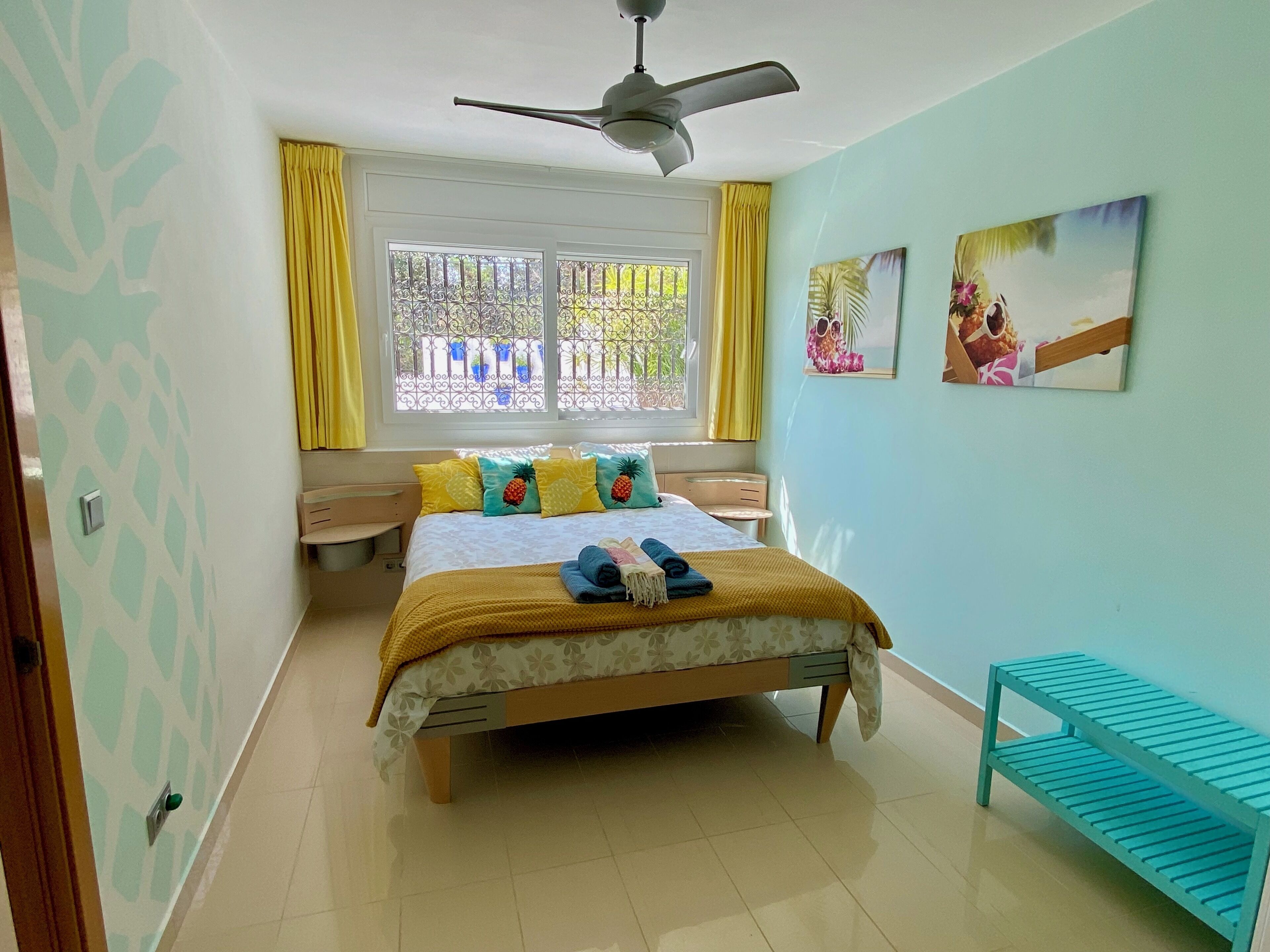 A bedroom with a bed, a ceiling fan, a window with yellow curtains, and a small table.