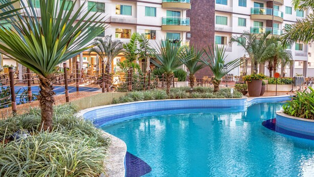 A pool area with palm trees, a tiled pool, and a building with balconies in the background. - Riviera Park Thermas - Oferta Caldas (Caldas Novas)