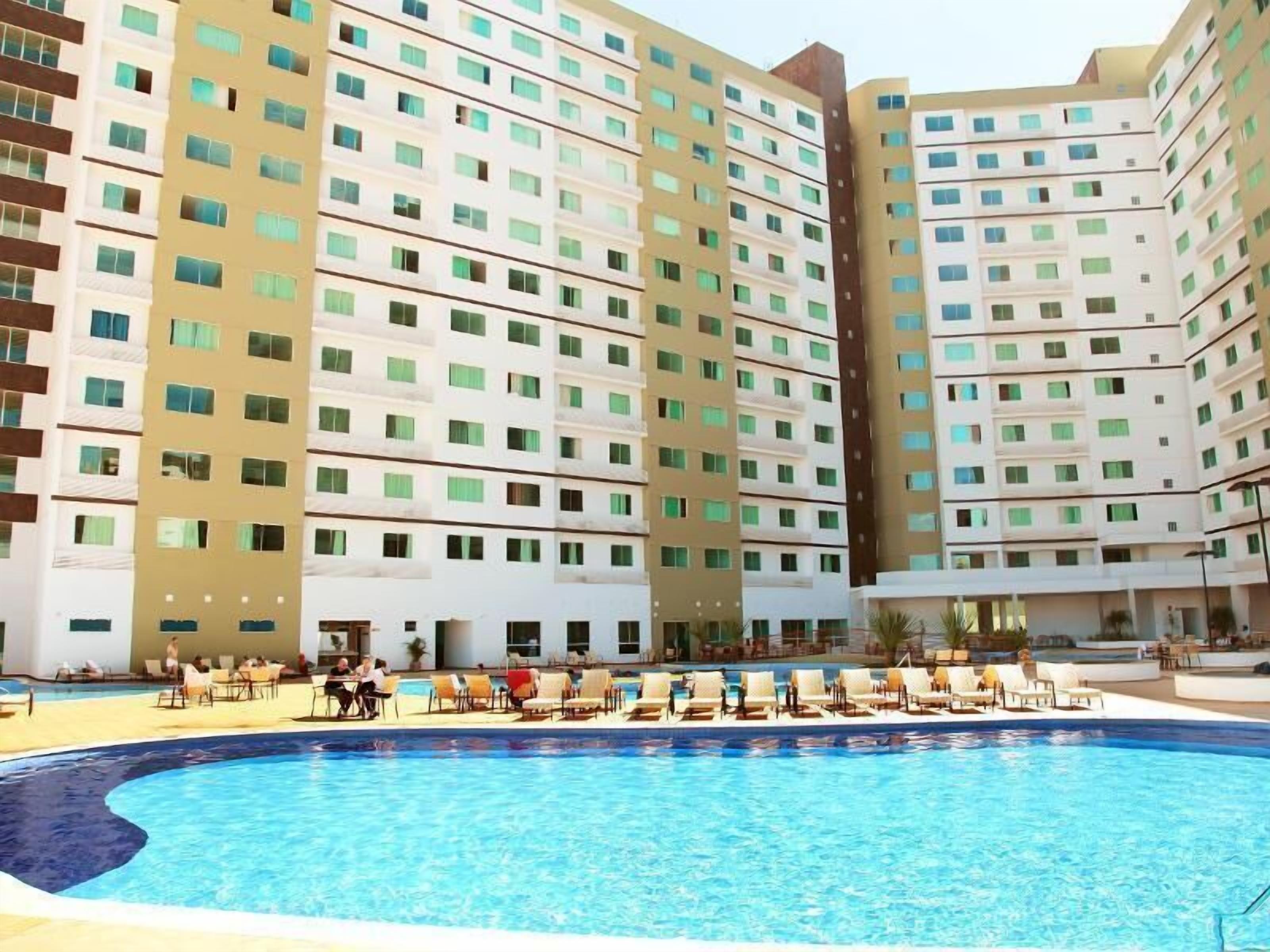 A hotel with multiple floors, a central pool area with lounge chairs, and a clear sky.