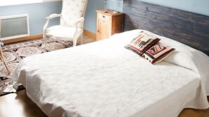 Een slaapkamer met een schuine balkenlaag, een bed met een witte dekbedovertrek, een stoel, een nachtkastje met een lamp, en twee schilderijen aan de muur.