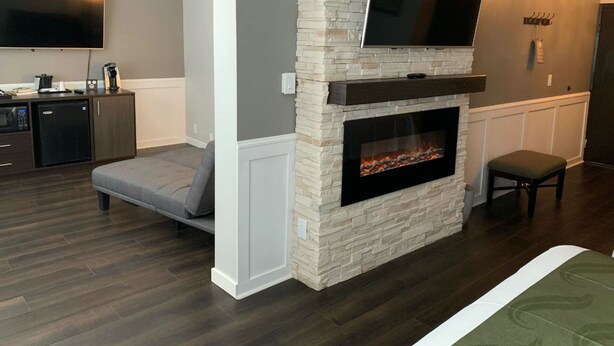A modern living room with a stone fireplace, a flat-screen TV mounted above it, a grey sofa, a small table, and a kitchen area with appliances. - Cedar Stables Inn and Suites (Sandusky)