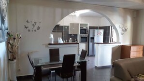 Une cuisine moderne dotée d’un îlot central, un coin repas avec une table et des chaises en verre, et un espace de vie comprenant un canapé et un meuble en bois.