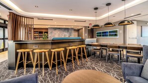 A modern hotel lobby with a curved reception desk, wooden bar stools, a seating area with tables and chairs, and a patterned carpet. - SpringHill Suites by Marriott Oakland Airport (Oakland)