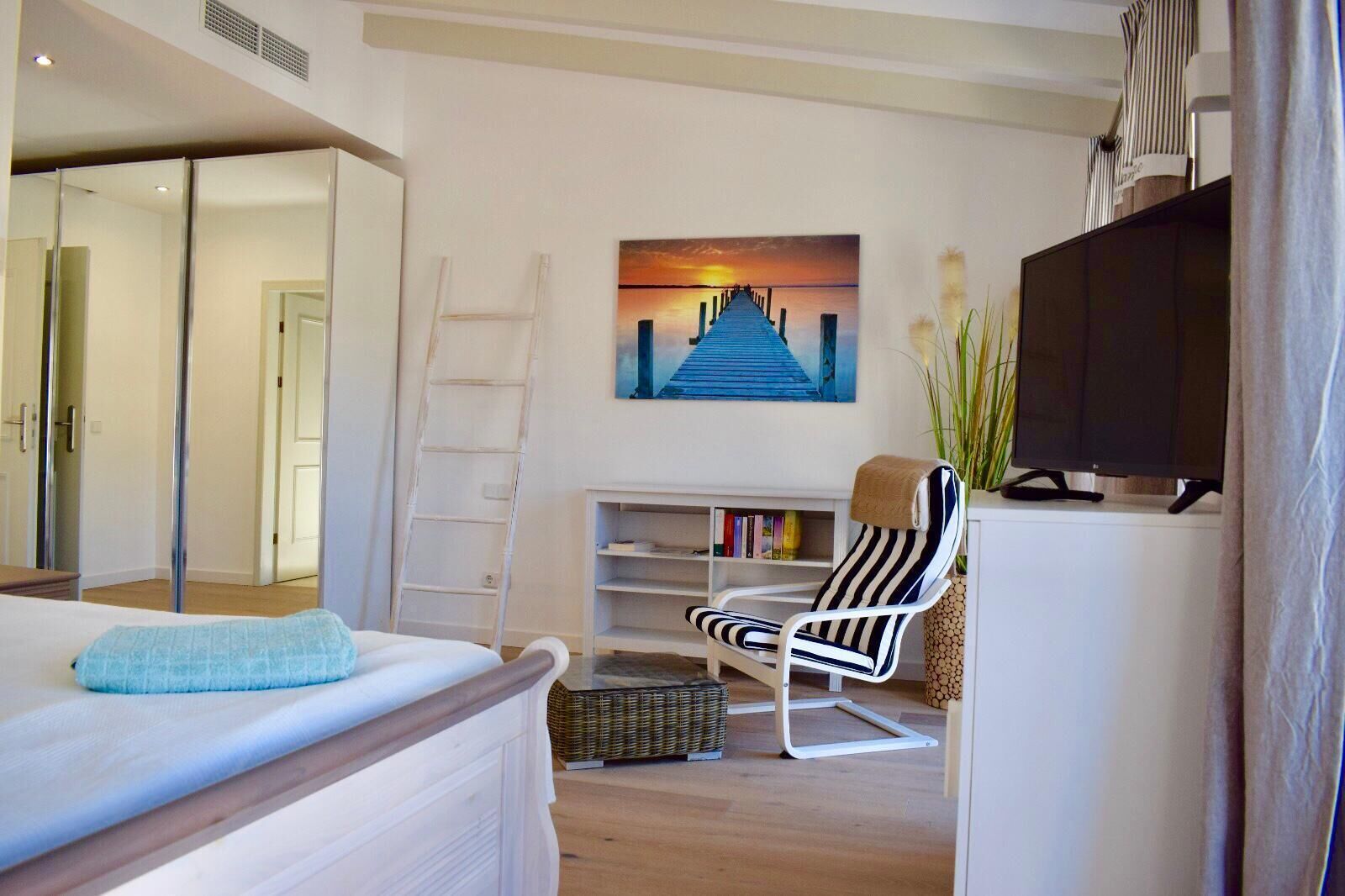 A modern bedroom with a bed, a chair, a TV, and a painting of a sunset over a pier.