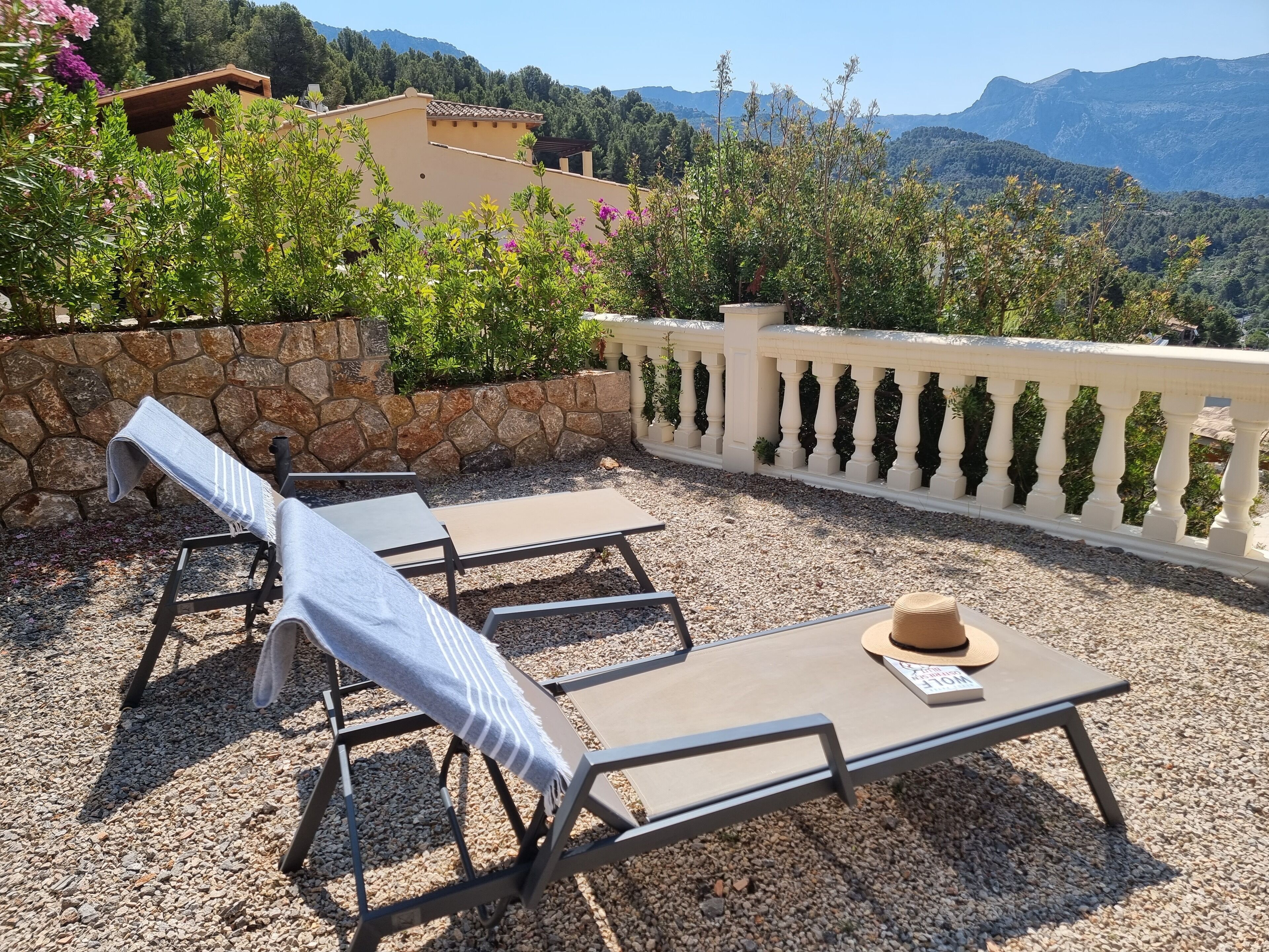 A terrace with lounge chairs and a table, surrounded by a stone wall and a white balustrade, overlooking a mountainous landscape.