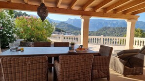 Un'area pranzo all'aperto riparata, con mobili in vimini, un tavolo imbandito con piatti e bevande, e una vista sulle montagne sullo sfondo.