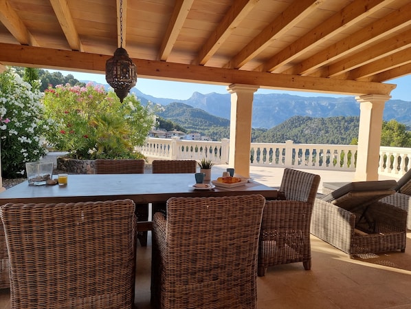A covered outdoor dining area with wicker furniture, a table with plates and a drink, and a view of mountains in the background.