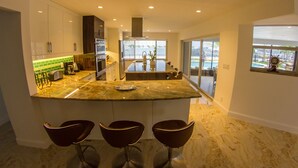 Cocina moderna con una isla central, tres taburetes de bar y vistas a una piscina a través de amplios ventanales.