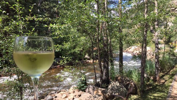 Una copa de vino blanco sobre un banco rojo, con vistas a un río, árboles y rocas.