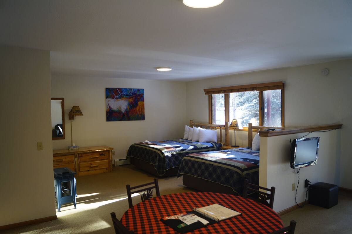 Ein Schlafzimmer mit zwei Betten, einem Schminktisch, einem Schreibtisch, einem Stuhl, einem Tisch mit kariertem Tischläufer, einem Gemälde, einem Fenster und einem Fernseher.