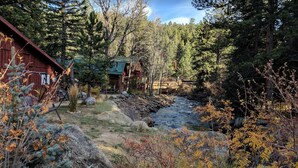 Una cabaña rústica frente a un río, rodeada de un bosque denso y follaje otoñal.