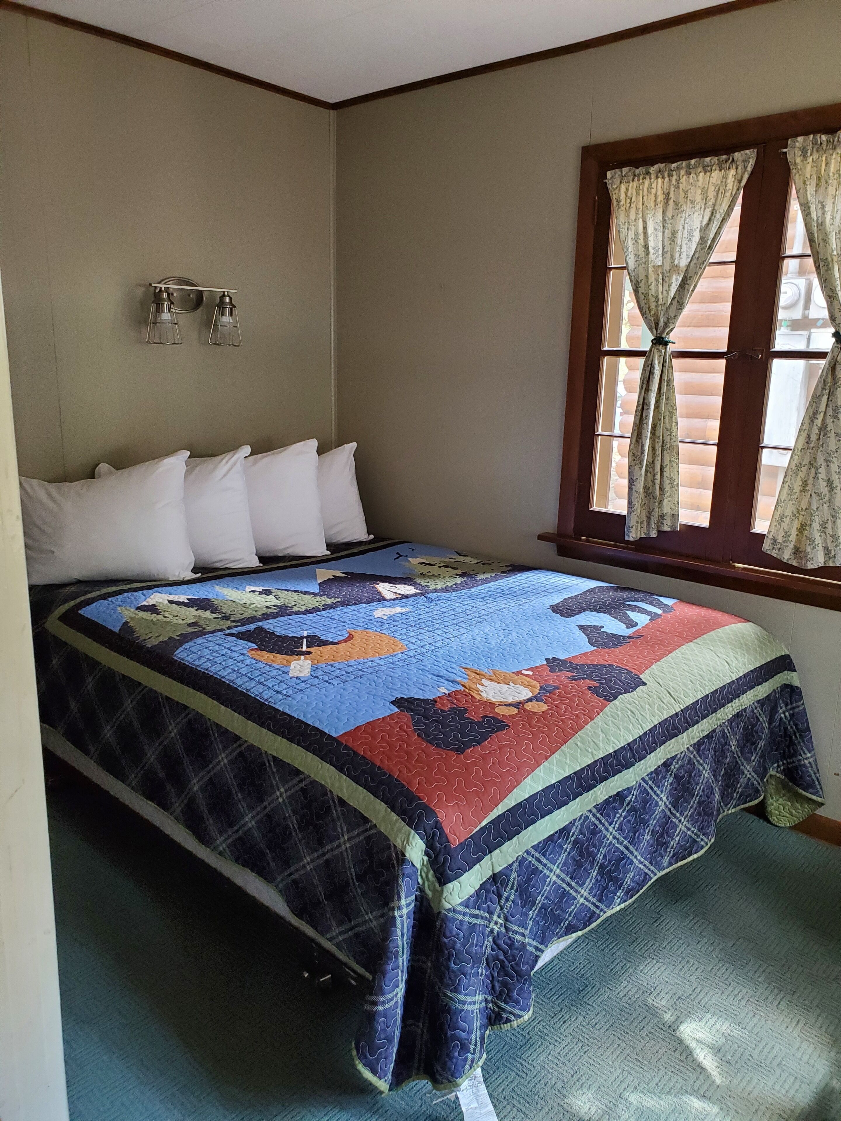 A bedroom with a bed, white pillows, a quilt, a window with curtains, and a wall-mounted light fixture.