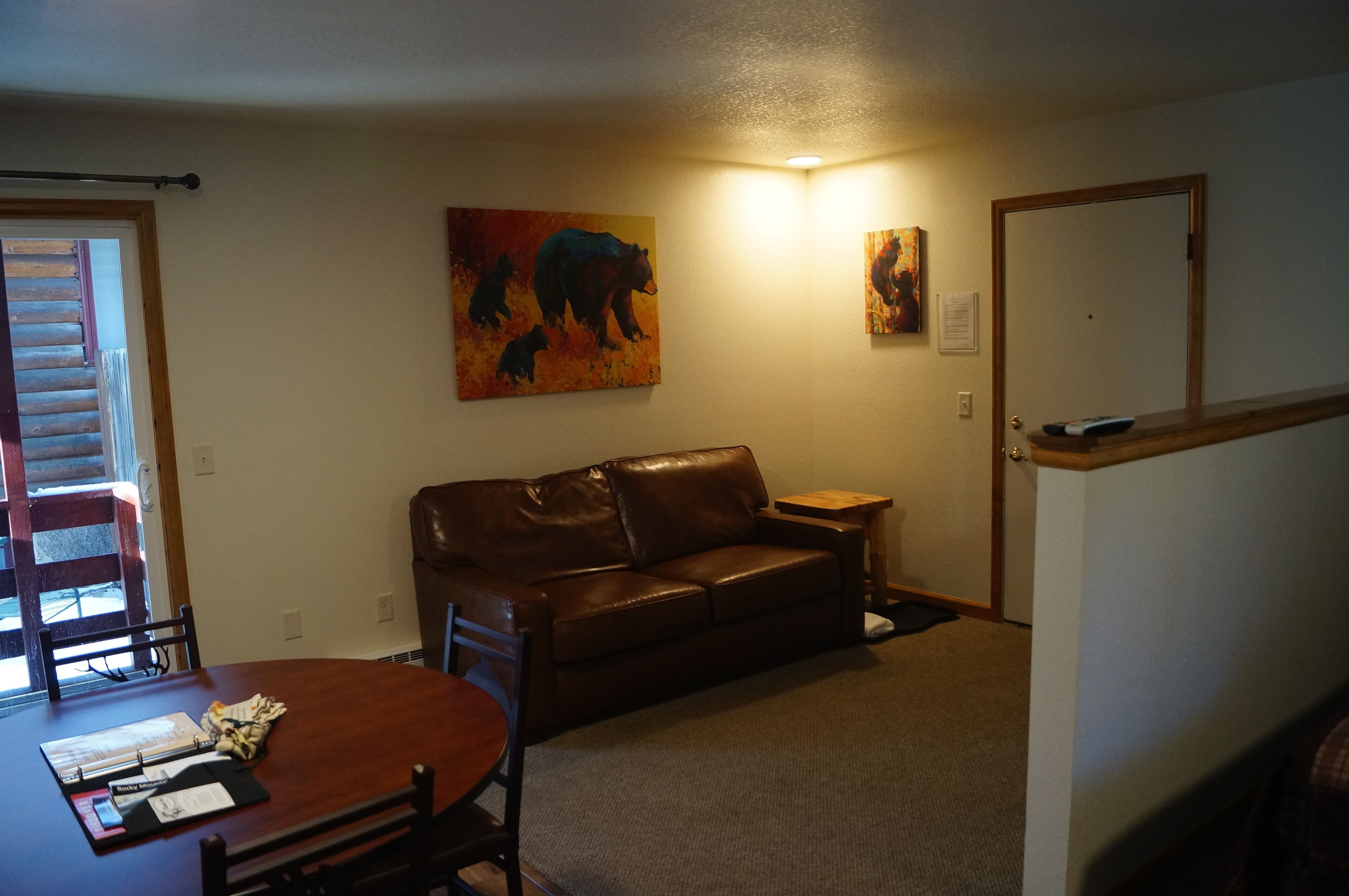 A living room with a brown leather sofa, a wooden coffee table, and a dining table with chairs. There are two paintings on the wall, a door, and a staircase.