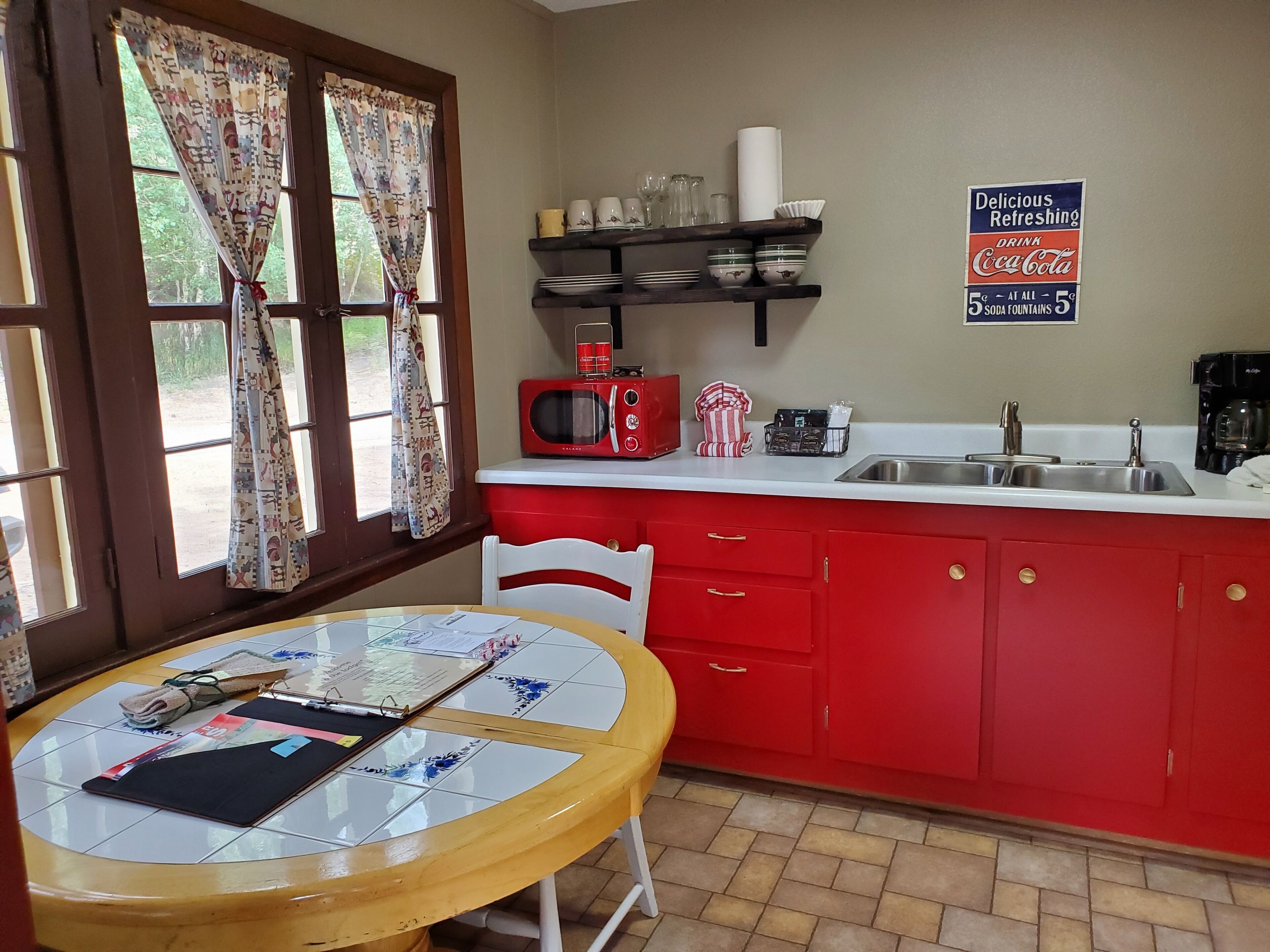 A kitchen with red cabinets, a white countertop, a red microwave, and a coffee maker. There is a window with curtains and a table with papers and a phone.
