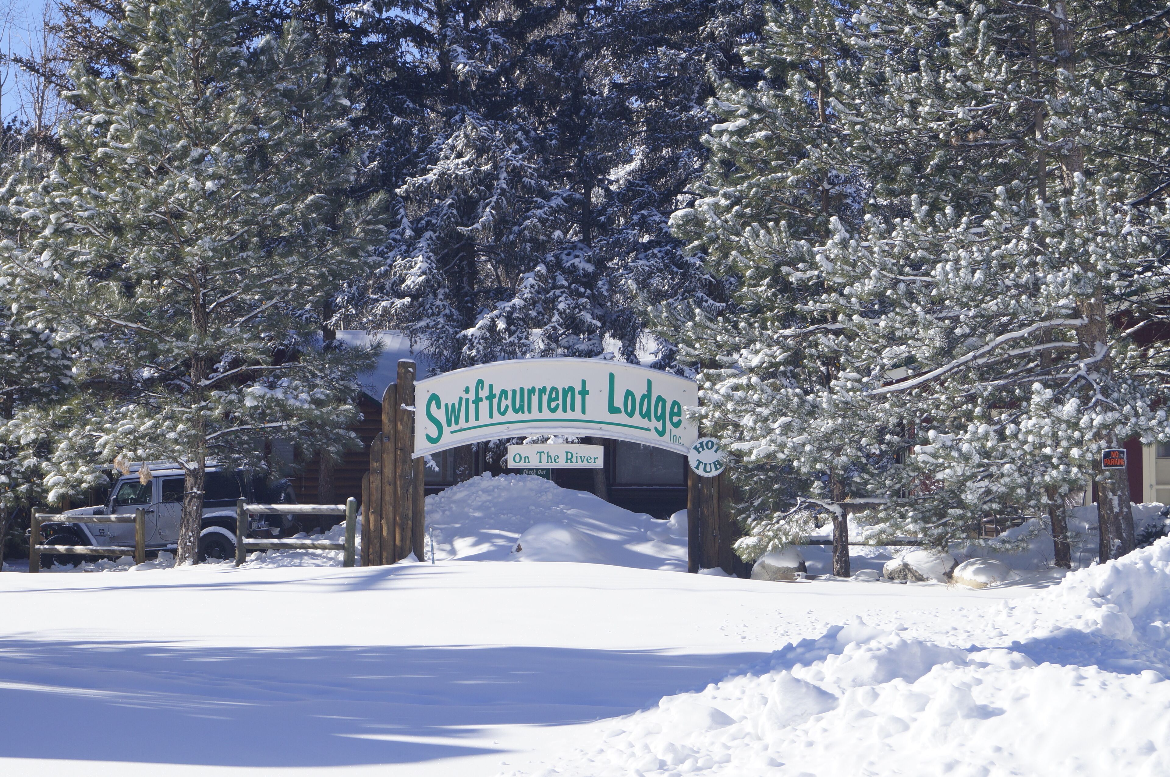 A snow-covered entrance to Swiftcurrent Lodge with a wooden sign and a Jeep parked nearby.