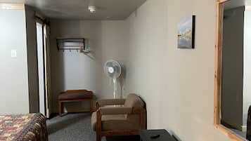Una camera d'albergo compatta con un letto, una sedia, un tavolino, una ventola portatile e una finestra.