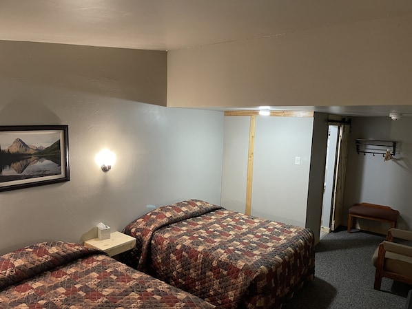 Habitación de hotel con dos camas, una mesita de noche, un cuadro enmarcado en la pared, un banco de madera y una pequeña mesa.