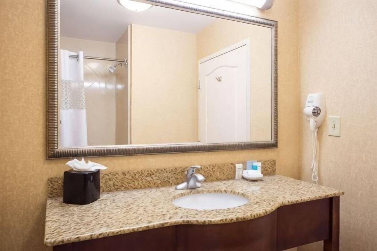 A bathroom with a large mirror, a sink, a towel dispenser, and a hairdryer on the wall.