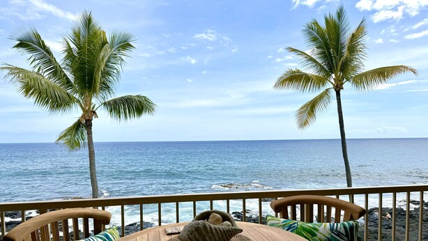 A wooden table with a woven placemat and a decorative shell, set against a backdrop of a clear blue sky and calm sea, with two palm trees framing the view.